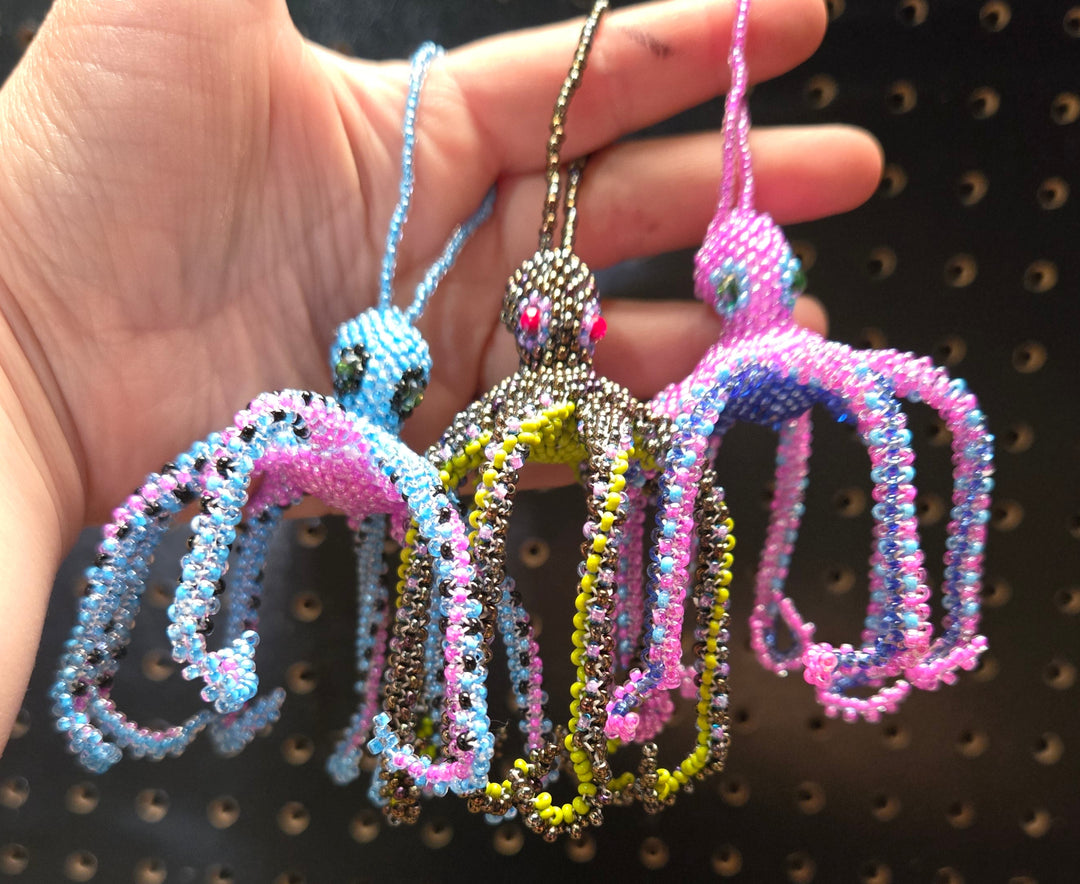 Three beaded octopus charms in pink, blue, and metallic silver, held in a person's hand against a black pegboard background.